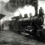 Authentic-style monochrome photo of a steam locomotive at a busy railway station platform in the 1880s.