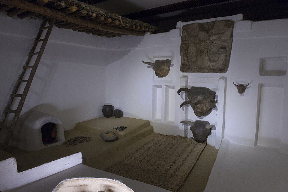 Reconstructed Neolithic house at Çatalhöyük with plastered walls, a hearth beneath the roof entry, and raised sleeping platforms