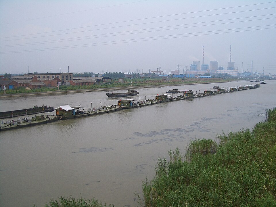 A modern industrial tugboat pushing a long line of flat steel barges down a wide river.