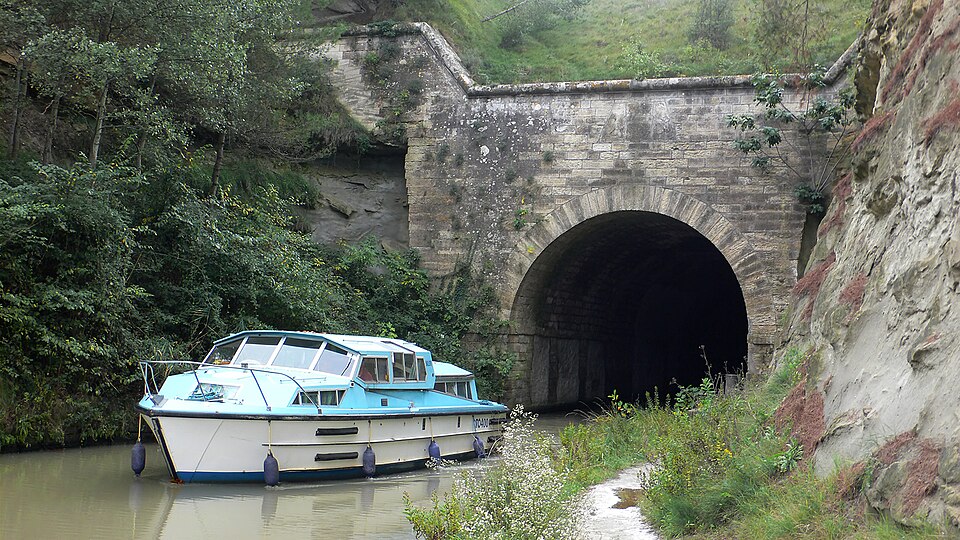 A canal barge emerging from a dark, arched stone tunnel carved through a hill.