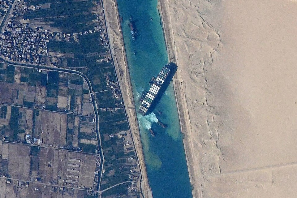 Satellite view of a massive cargo ship wedged diagonally across a narrow desert canal.